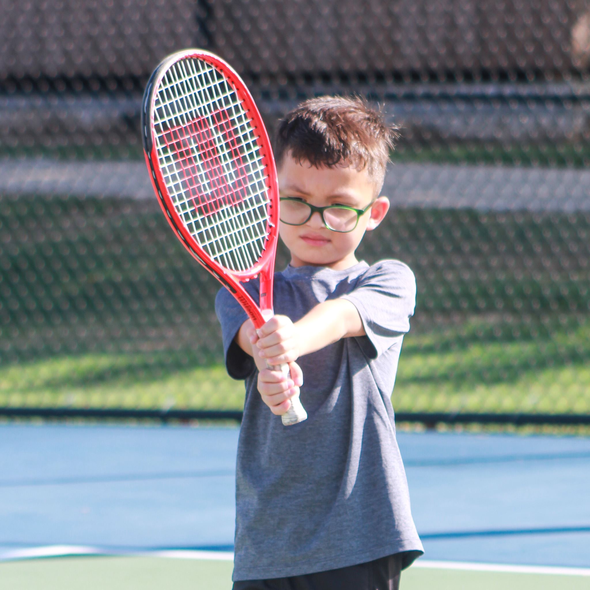 Young boy in glasses with arms extended holding a tennis racket